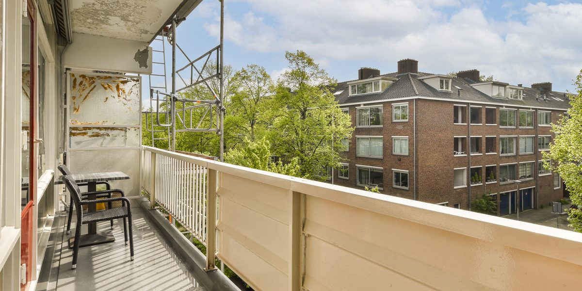 Apartment balcony with a railing and chairs, facing trees and buildings across the street